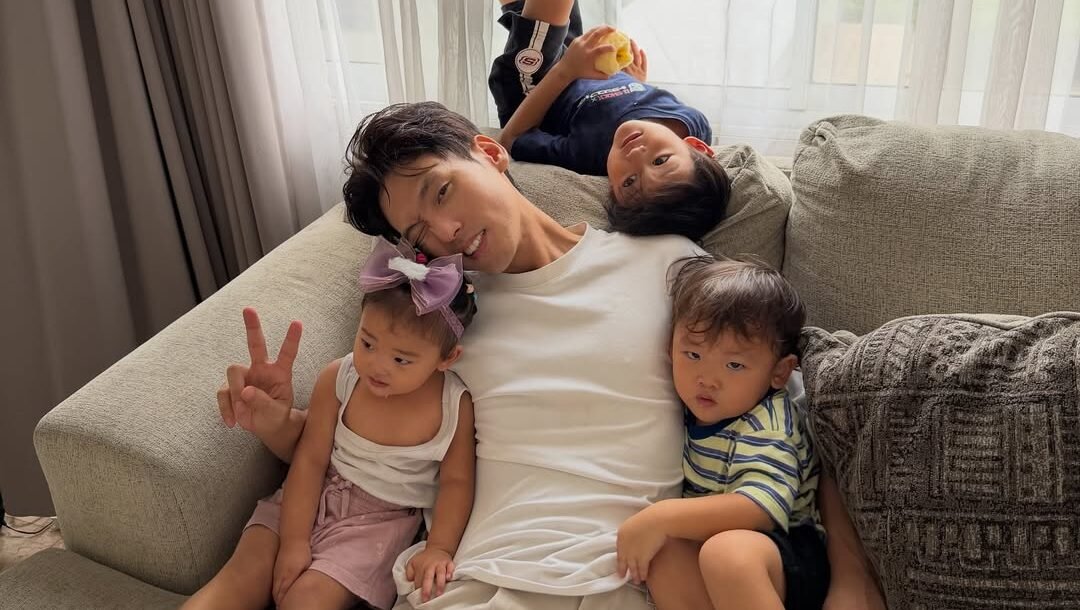 Daehoon yang terlihat bermain dan bercengkerama hangat bersama dua anak di ruang keluarga, menggambarkan keterlibatan ayah dalam pengasuhan meski ada bantuan babysitter.